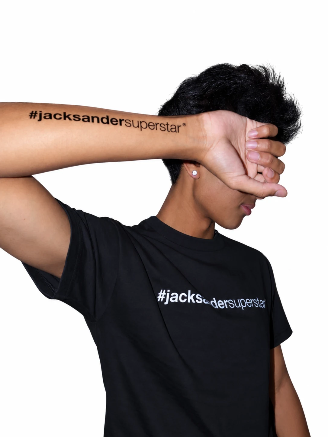 Model in a black jacksandersuperstar tee with a raised forearm across the face, showing matching lettering.